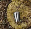 A stack of metal cups on a rough wooden surface, with a blurred forest floor in the background. Náhled obrázku produktu