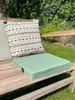A close-up of a green outdoor seat cushion and a patterned throw pillow on a wooden bench, with lush green foliage in the background. Produktbild-Vorschau
