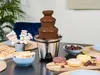 A chocolate fountain with various dipping items like marshmallows, donuts, and cookies, set on a table. Podgląd zdjęcia produktu