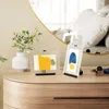 Two abstract art prints in frames are displayed on a wooden dresser next to a white vase with greenery. A large, gold-rimmed mirror reflects a bed in the background. Podgląd zdjęcia produktu
