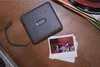 A gray Fujifilm Instax Link Wide printer is shown on a wooden surface next to a stack of printed photos. Produktbild-Vorschau