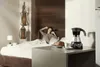 A woman is stretching in bed while a De'Longhi Moka pot, a croissant, and a cup of coffee are on a table in the foreground. Produktbild-Vorschau