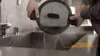 A person is rinsing a pot with a lid under a faucet in a kitchen sink. Podgląd zdjęcia produktu
