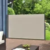 A retractable side awning is extended on a wooden deck, providing privacy and shade. The awning is beige and has gray metal posts. In the background, there are green trees and a lawn. Produktbild-Vorschau
