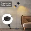 A black floor lamp with two adjustable heads is shown in a living room setting next to a sofa. Produktbild-Vorschau
