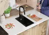 A modern kitchen island with a black sink, a cutting board with ingredients, and a person holding a coffee cup. Produktbild-Vorschau
