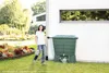 A woman is watering plants next to a green rain barrel. Voir les images du produit