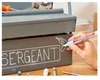 A hand is using a white marker to draw on a toolbox. The toolbox has the word "SERGEANT" written on it in white chalk-like lettering, and a drawing of a drill is being added next to it. Tools are visible in the background. Produktbild-Vorschau
