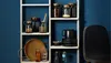 A white shelving unit filled with various kitchen items, including jars, bowls, and a tray, against a dark blue wall. Produktbild-Vorschau