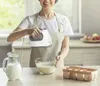 A woman is using a white and gray hand mixer to mix ingredients in a clear glass bowl. Podgląd zdjęcia produktu