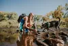 A woman and her dog are in a shallow river, with the woman filling a water bottle. Produktbild-Vorschau