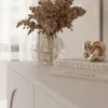 A close-up of decorative items on a white surface, including a vase with dried flowers, books, and a metallic knot sculpture. Produktbild-Vorschau