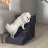 A small white dog is climbing on a dark gray pet stair next to a sofa. Produktbild-Vorschau