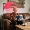 An elderly man is sitting on a leather couch reading a book, with a red light therapy panel shining on him from a stand. Produktbild-Vorschau