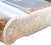 A close-up view of a clear plastic food storage container filled with small brown particles, likely food items like grains or seeds. The container has some embossed markings on its side. Produktbild-Vorschau