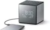 A cube-shaped digital alarm clock with a USB port connected to a smartphone. Produktbild-Vorschau