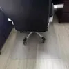 The back of a black office chair is shown on a clear plastic floor mat on a light wood-grain floor. The chair has silver legs and black wheels. Náhled obrázku produktu