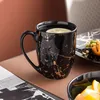 A black mug with gold marbling, filled with water and a lemon slice, sits on a brown surface next to a plate of food and a magazine. Náhľad obrázku produktu
