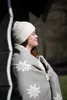 A woman wearing a white beanie and wrapped in a gray blanket with white edelweiss patterns leans against a dark wooden structure. Produktbild-Vorschau
