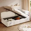 A beige upholstered bed with its storage compartment open, revealing stored items. The bed is in a room with a wooden floor and a window with curtains. Anteprima dell’immagine prodotto