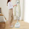 A woman is using a garment steamer on a white shirt in a room with a bed and a window. Produktbild-Vorschau