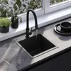 A black kitchen sink with a black faucet is shown with water running into it. The sink is on a gray countertop next to a window with white flowers outside. There are two small potted plants on the windowsill. Náhled obrázku produktu