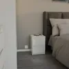 A minimalist white nightstand with two drawers is shown next to a bed in a bedroom setting. Voir les images du produit