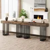 A long, narrow console table with a rustic wood top and a black metal slatted base, placed behind a sofa in a living room setting. A fireplace with a fire is visible in the background. Náhľad obrázku produktu