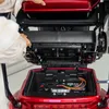 A person's hands are shown opening the battery compartment of a red mobility scooter. Produktbild-Vorschau