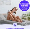 A woman is sitting on a bed, wearing headphones and smiling. Produktbild-Vorschau