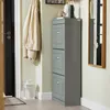 A narrow shoe cabinet with three flip drawers stands in a hallway next to a door with a coat hanging on it. A mirror is visible on the right wall. Podgląd zdjęcia produktu