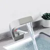 A modern silver waterfall faucet is shown in use, with water flowing into a sink. A small potted plant is visible in the background. Podgląd zdjęcia produktu