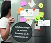 A woman is peeking around a refrigerator door that is covered in notes, magnets, and a check. The notes include a drawing of a voucher, a smiley face, and a "Good morning" message. Produktbild-Vorschau