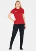 A woman is posing in a studio setting wearing a red t-shirt and black pants. Produktbild-Vorschau