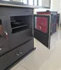 A close-up view of a black wood-burning stove with an open oven door, revealing a red interior and a pan inside the oven. Another stove is visible in the background. Produktbild-Vorschau