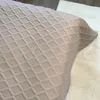A close-up view of a taupe bedspread with a diamond pattern, draped over a bed and a fluffy rug. Podgląd zdjęcia produktu