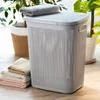 A laundry basket with a lid, placed on a wooden floor next to a stack of folded towels and a potted plant. Produktbild-Vorschau