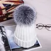 Weiße Strickmütze mit großem flauschigen grauen Pompon oben, auf einem aufgeschlagenen Magazin neben einer Brille liegend Produktbild-Vorschau