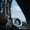 A G-SHOCK watch is placed on a dark surface with a blurred background of a bridge and sky. Náhled obrázku produktu