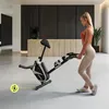 A woman is standing next to a stationary bike in a modern home setting. Produktbild-Vorschau