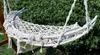 A white macrame hammock chair is suspended outdoors against a backdrop of green foliage. Produktbild-Vorschau