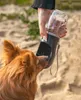 A person is holding a dog water bottle for a dog to drink from on a sandy beach. Produktbild-Vorschau