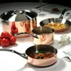 A collection of copper cookware, including a large pot with a lid, a saucepan, and a frying pan, arranged on a white tablecloth with some tomatoes and a flower. Produktbild-Vorschau