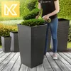 A person is watering plants in large, square, textured planters on a wooden deck, with a lush green hedge in the background. Produktbild-Vorschau