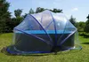 A round swimming pool covered by a transparent dome-shaped tent with blue trim, set up on a grassy lawn with trees in the background. Produktbild-Vorschau