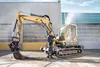 A person is pressure washing a yellow excavator in front of a building. Produktbild-Vorschau