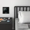 A smart thermostat is mounted on a wall next to a bed with a nightstand containing a phone and charger. Náhled obrázku produktu