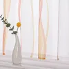 A close-up of a sheer curtain with vertical wavy stripes in orange, pink, and beige, with a small vase holding a single yellow flower and a sprig of greenery in the foreground. Produktbild-Vorschau
