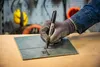 A gloved hand holding a Sharpie marker is marking a metal plate on a wooden surface. The background shows tools and safety equipment. Anteprima dell’immagine prodotto