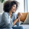 A woman is sitting on a couch, looking to the side and smiling. She is wearing a smartwatch on her wrist. Voir les images du produit